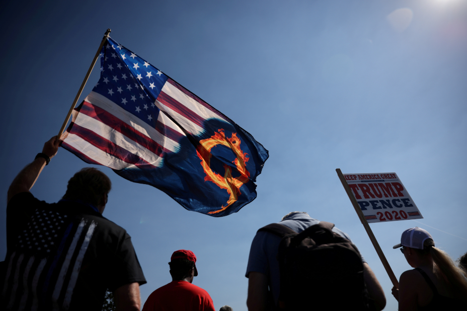 photo of man at rally holding us flag emblazoned with burning letter Q