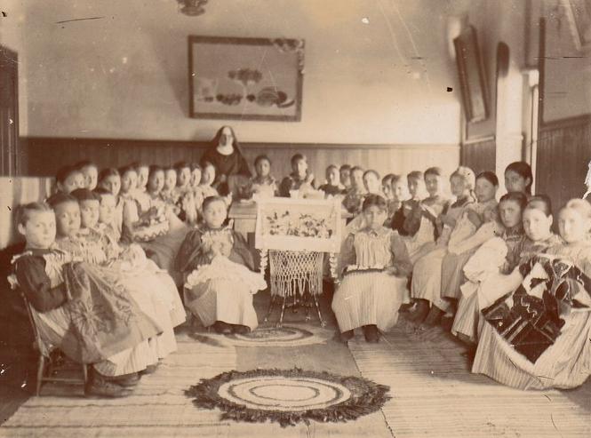 Photo of girls showing their handiwork in the work room at St. Benedict's Mission School, circa 1895.